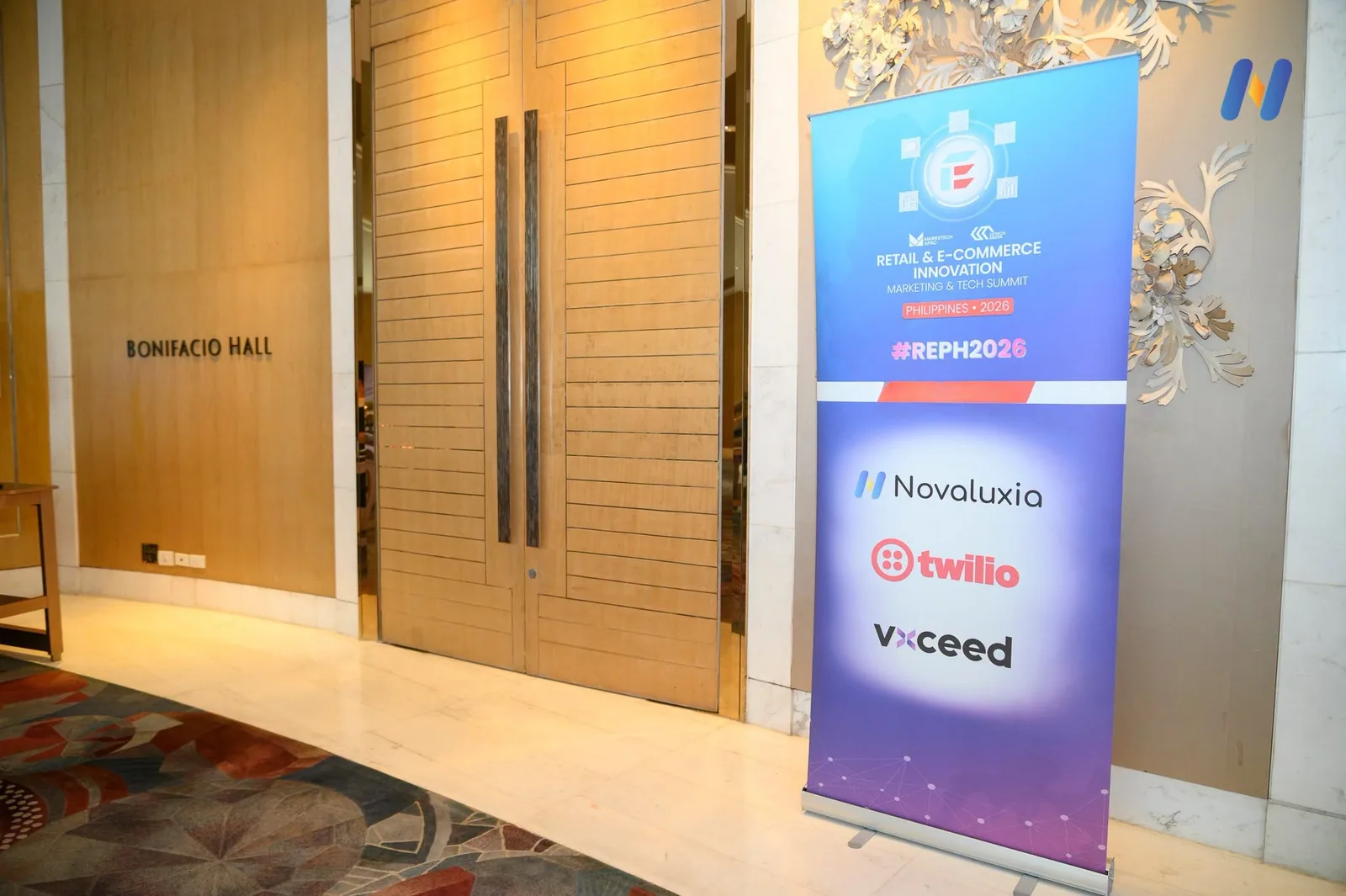 Novaluxia PH listed as a sponsor on the Retail and E-Commerce Innovation Summit Philippines 2026 board outside Bonifacio Hall at Shangri-La The Fort
