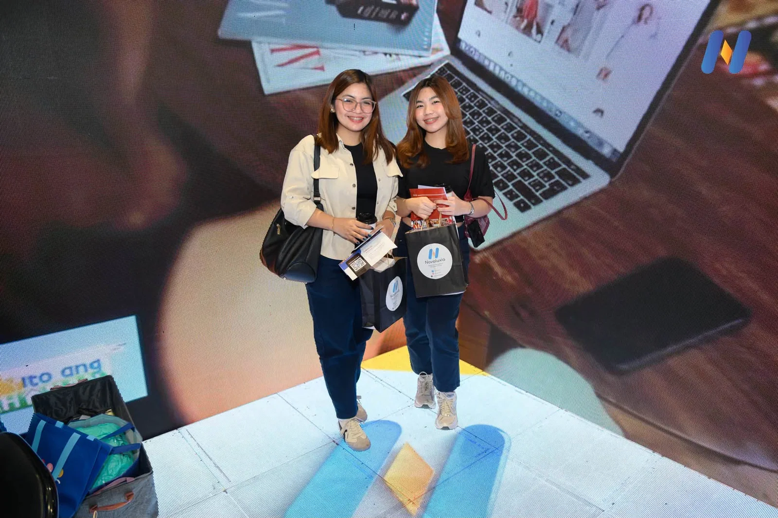 Two visitors holding Novaluxia tote bags and brochures in front of the platform's app display wall at the expo booth